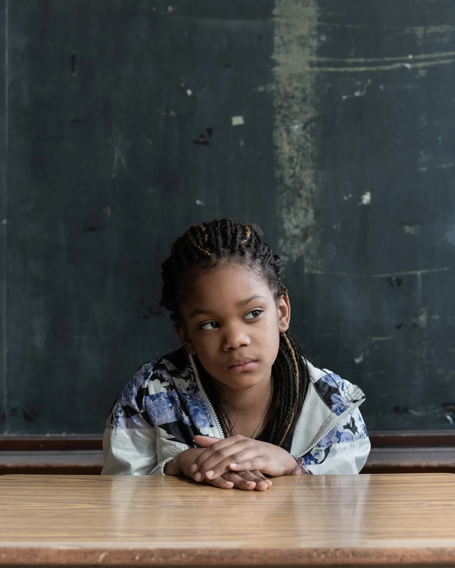 A portrait of a young person, perhaps a third to fifth-grader, with their head tilted and eyes looking to their left, hands laying flat on a table in front of them, one on top of the other. behind them is well-worn black chalkboard.