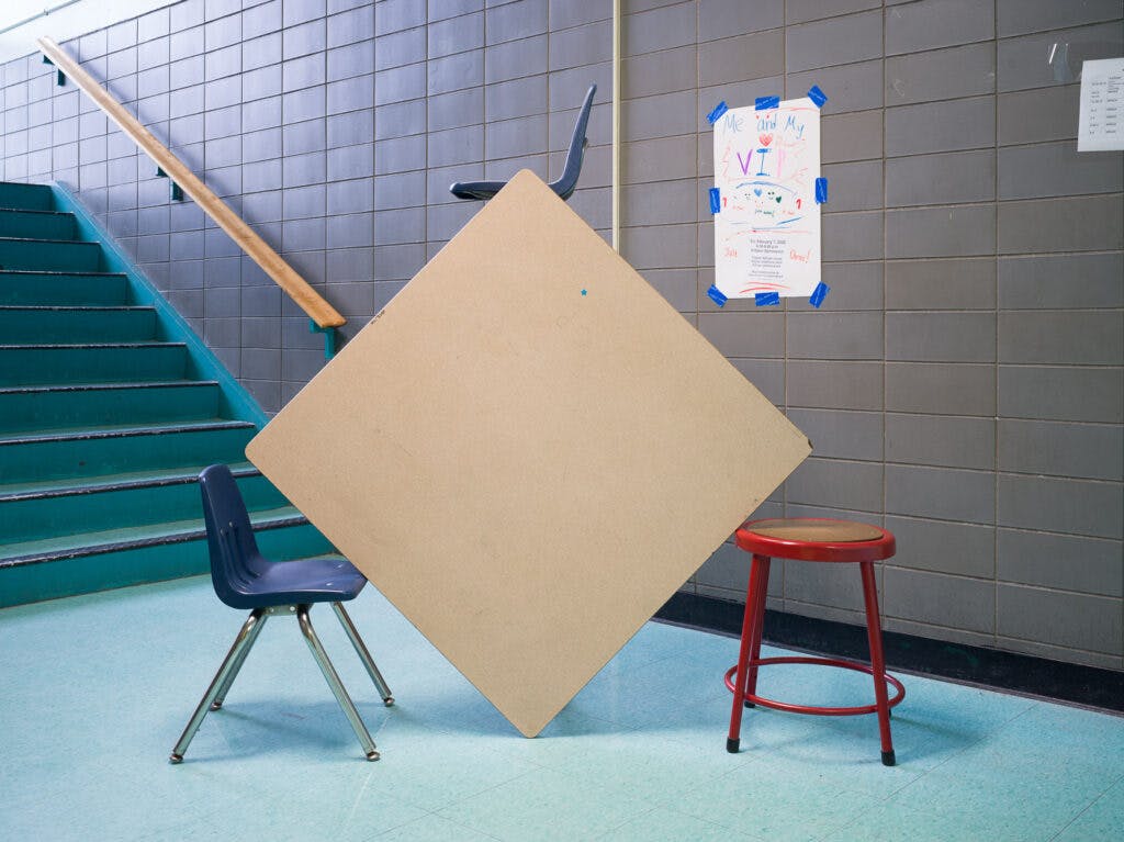 A photo of a grey school table, flipped on its side to form the shape of a diamond, supported by red & blue chairs on each side. a blue chair peeks up from the top corner. there's a blue stairway rear-left and a crayon poster on the wall behind